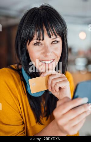Glückliche, reife Frau mit Kopfhörern um den Hals, die mit Kreditkarte und Mobiltelefon verwendet wird Stockfoto