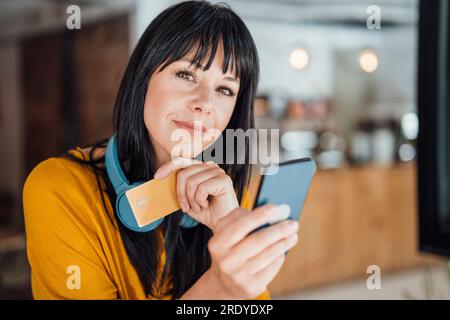 Lächelnde Frau mit Kopfhörern um den Hals in der Hand eines Mobiltelefons und einer Kreditkarte im Café Stockfoto