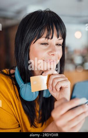 Lächelnde, reife Frau mit Kopfhörern um den Hals, die Mobiltelefon und Kreditkarte nutzt Stockfoto