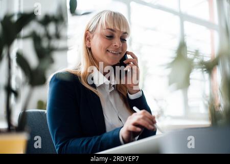 Glückliche Geschäftsfrau, die einen Tablet-PC verwendet und im Büro mit einem Smartphone spricht Stockfoto