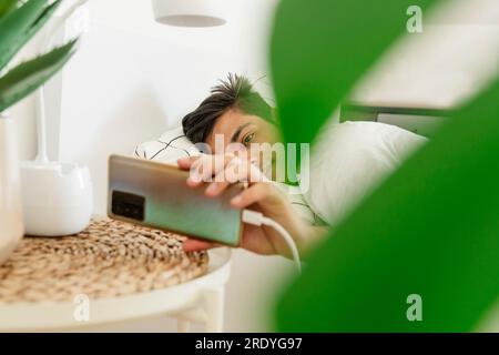 Lächelnder Mann, der sein Smartphone in der Nähe des Bettes auflädt Stockfoto