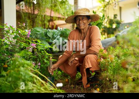 Ältere indische Farmerin, die Blumen pflanzt und glücklich lächelt. Ältere Sri-lankische fröhliche Frau, die in ihrem Garten sitzt und sich um blühendes Grün kümmert Stockfoto