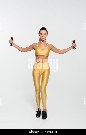 Eine Frau in Fitnesskleidung trainiert mit Kurzhanteln und schaut auf weißem Hintergrund in die Kamera Stockfoto