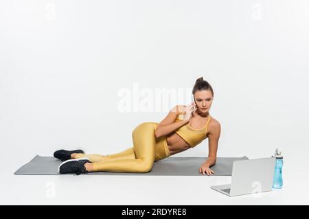 Frau, die mit einem Smartphone spricht, in der Nähe eines Laptops sitzt und eine Sportflasche auf einer Fitnessmatte hat, weißer Hintergrund Stockfoto