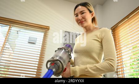 Junge Frau, die einen kabellosen Handstaubsauger verwendet, um den Staub im Wohnzimmer zu saugen und zu reinigen Stockfoto