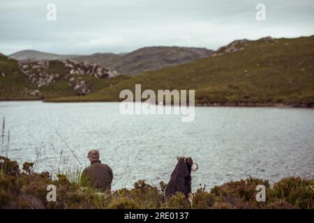 Mann und Hund sitzen Fliegenfischen am loch in abgelegenen Bergen in schottland Stockfoto