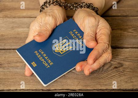 Der biometrische Reisepass der Ukraine wird in den Händen einer älteren Frau gehalten, deren Hände mit einer Kette auf einem Holzbrett gebunden sind, Einschränkungen für ein Ukra Stockfoto
