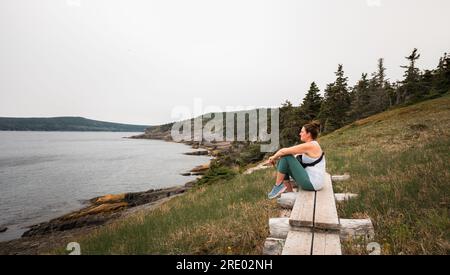 Eine Frau sitzt auf einem hölzernen Pfad und sieht auf einer Wanderung aufs Meer. Stockfoto