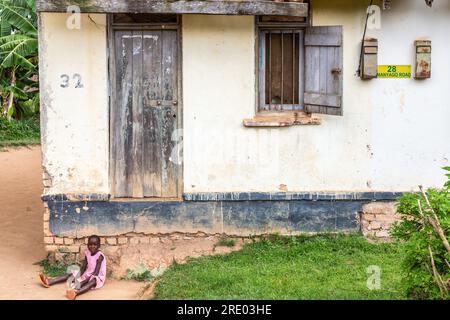 Sehr bescheidenes Wohnhaus in einer Straße in Entebbe, Uganda. Ein junges Mädchen in Rosa, das an der Türschwelle sitzt. Stockfoto