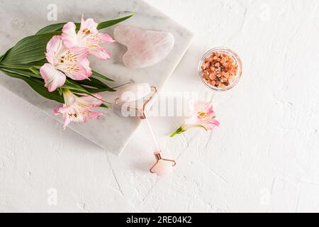 Draufsicht auf die Massagewalze aus Rosenquarz, Gouache, Salz in einer Schüssel auf weißem Hintergrund mit Aromeria und Marmorstein. Hautpflege Stockfoto