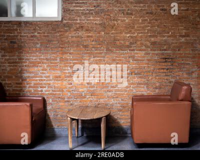 Braunes, luxuriöses Einzelsofa aus modernem Leder, Seitenblick mit rundem Holzbeistelltisch, dekoriert auf leerem Backsteinhintergrund. Innenausstattung im Loft-Stil mit Stockfoto