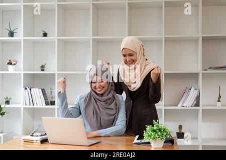 Zwei junge muslimische Frauen, die in Meetings arbeiten und darüber diskutieren, freuen sich über gute Nachrichten im Konferenzraum mit Finanzdiagrammen und Laptop Stockfoto