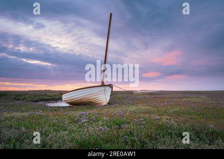 Ein kleines Segelboot inmitten des Lavendelmeers in den Salzmarschen von Brancaster Staithe bei Sonnenuntergang. Stockfoto