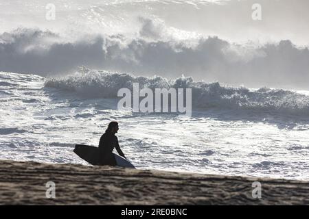 Silhouette eines männlichen Surfers, der am Strand vor dem Hintergrund großer Wellen spaziert Stockfoto