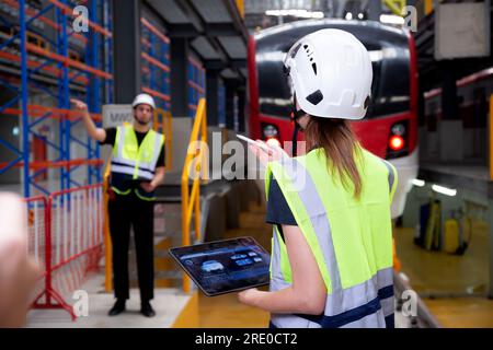 Junge, weiße Ingenieurin, die ein digitales Tablet benutzt, und ein Mann oder Arbeiter, der den Elektrozug für die Planung von Wartungsarbeiten in der Station, im Transport und in der Infrastruktur überprüft Stockfoto