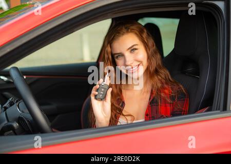 Mit einem fröhlichen Lächeln sitzt eine rothaarige Frau in einem karierten Hemd bequem in einem brandneuen roten, modernen Auto und hält die Autoschlüssel freudig fest. Stockfoto