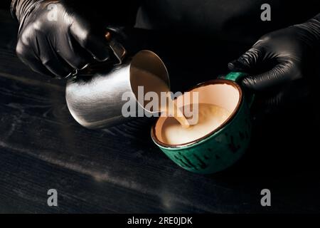 Teilweise, Barista in Latexhandschuhen, der Cappuccino, Espresso, Latte Art, Pitcher mit Milch zubereitet Stockfoto