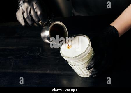 Barista in Latexhandschuhen bereitet Latte Macchiato zu, gießt Espresso ein, Pitcher mit schwarzem Kaffee Stockfoto