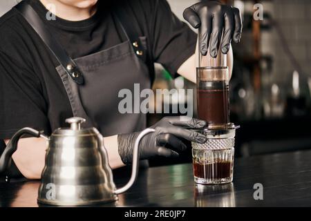 Teilansicht des Baristas in schwarzen Handschuhen und Schürze beim Zubereiten des Espressos in der Aero Press in der Nähe des Wasserkochers Stockfoto