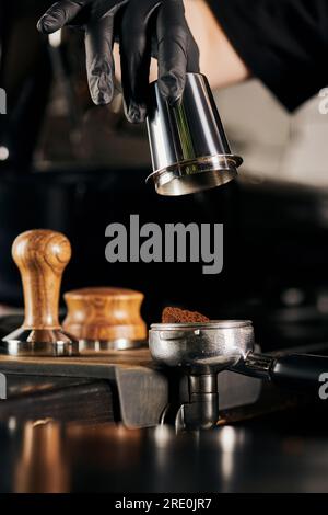 Zugeschnittene Ansicht des Baristas in schwarzem Latexhandschuh, der den Messbecher in der Nähe des Portafilters hält Stockfoto