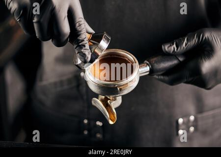 Zugeschnittene Ansicht von Manipulationsgerät mit gemahlenem Kaffee und Portafilter in den Händen des Baristas in schwarzen Handschuhen Stockfoto