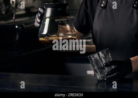 Der Barista in schwarzen Handschuhen hält Glas und Kaffeekanne mit frisch gebrühtem aromatischem Espresso Stockfoto