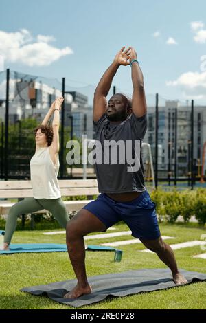 Junger, aktiver afroamerikanischer Mann in Sportbekleidung, der auf einer Matte auf einem Sportplatz steht und beim Sport die Arme über dem Kopf hält Stockfoto