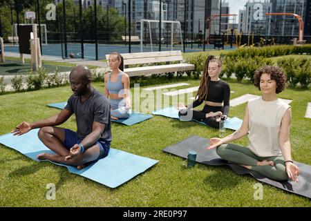 Gruppe junger interkultureller Menschen in Aktivkleidung, die in Lotusposition auf Matten sitzen und Yoga auf dem grünen Rasen des Sportplatzes üben Stockfoto