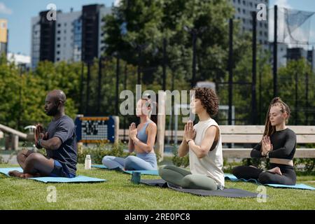 Seitenansicht einer Gruppe interkultureller Athleten, die Yoga im Freien üben, während sie auf Matten sitzen, mit überkreuzten Beinen und Handflächen an der Brust zusammengelegt Stockfoto