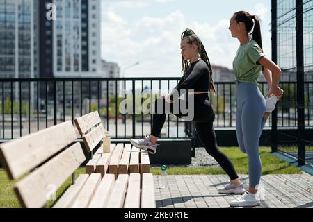 Zwei junge aktive Frauen in Sportbekleidung stehen an sonnigen Tagen auf einer Bank und trainieren die Beinmuskulatur im Freien Stockfoto