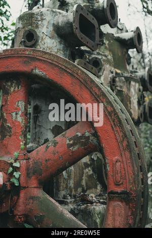 Detail des roten Rads eines alten verrosteten Luftkompressors im Wald. Industrielle Geschichtsmaschine. Eisen- und Stahlproduktion. Verlassene Maschinen. Stockfoto