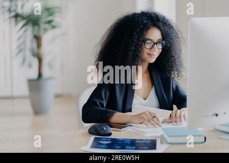 Remote Afro Freiberufler, auf Schreiben fokussiert, freudiger Ausdruck, geräumiger Schrankarbeitsplatz, Topfpflanze. E-Learning-Konzept. Stockfoto