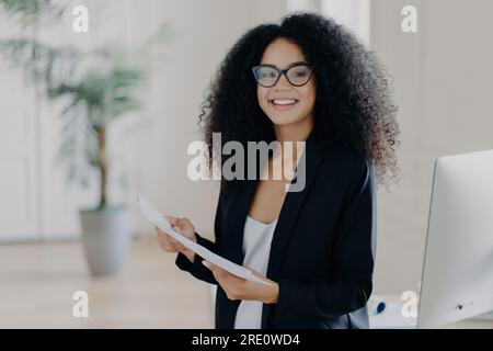 Fröhliche Afro-Unternehmerin in eleganter Kleidung, Brille tragen, Dokumentation studieren, im Büro stehen, sich auf die Präsentation von Geschäftsideen vorbereiten Stockfoto