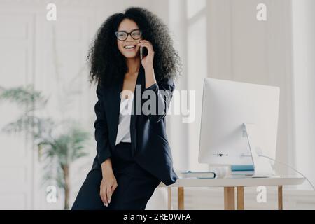 Zufriedene dunkelhäutige Geschäftsfrau in der Pause, die sich auf einen weißen Tisch lehnt, mit einem Computer im Büro, mit ihrem Handy spricht, eine Brille und ein Ei trägt Stockfoto