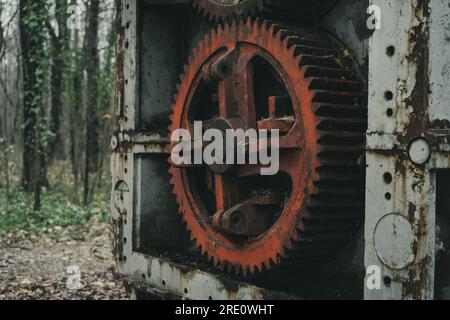 Nahaufnahme des roten Zahnrads einer alten verrosteten, verrosteten Blechbearbeitungsmaschine im Wald. Eisen- und Stahlproduktion. Industrielle Geschichtsmaschine. Stockfoto