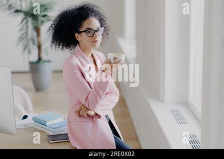 Lockige Frau, aufmerksam, hält Kaffee, blickt aus dem Fenster in den Gemeinschaftsraum im Büro. Stockfoto