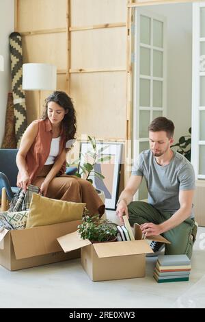 Junger Mann und Frau, die Kartons mit Büchern, grünen Hauspflanzen und Bettwäsche packen, während sie in eine neue Wohnung ziehen Stockfoto