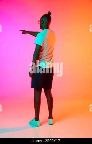 Bild in voller Länge von einem afrikaner mit Brille, der seine Hand nach oben hält und etwas auf einen orange-rosa Hintergrund in Neon zeigt. Stockfoto