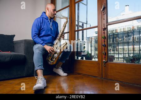 Junger venezolanischer Latino-Mann, Saxophonist spielt Inspiration im Wohnzimmer seiner Wohnung, sitzt auf dem Sofa neben dem Balkonfenster. Stockfoto