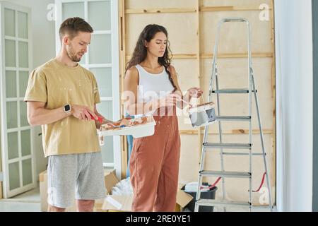 Ein junges Paar in Freizeitkleidung hält Plastikeimer und -Tablett und mischt Farben, bevor es Wände des geräumigen Wohnzimmers einer neuen Wohnung streicht Stockfoto
