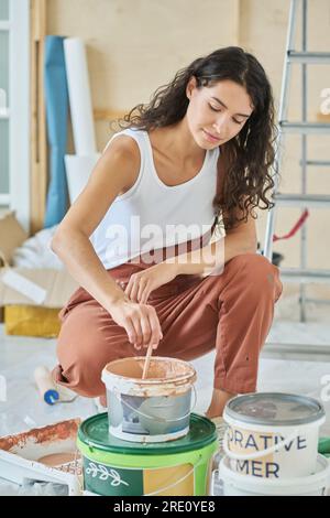 Junge Frau in Overalls und weißer Baumwoll-Weste, die auf Kniebeugen vor der Kamera sitzt und Farbe mischt, während sie sich auf das Auftragen an der Wand vorbereitet Stockfoto