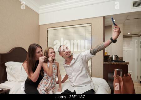 Glückliche Mutter und Tochter, die mit den Fingern eine Herzgeste machen, wenn der Vater Selfie mit ihnen nimmt Stockfoto