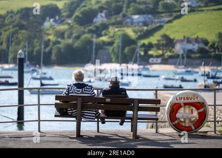 Das Paar sitzt auf einer öffentlichen Bank auf dem Whitestrand Parkplatz, im Zentrum von Salcombe, Devon, mit Blick auf die angelegten Boote im Hafen an einem sonnigen Tag Stockfoto