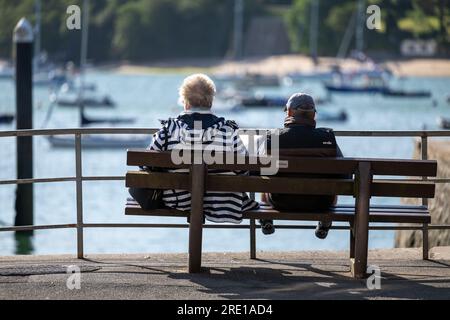 Das Paar sitzt auf einer öffentlichen Bank auf dem Whitestrand Parkplatz, im Zentrum von Salcombe, Devon, mit Blick auf die angelegten Boote im Hafen an einem sonnigen Tag Stockfoto