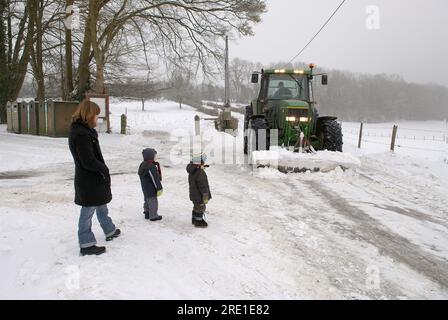 Bauer, der Schnee von einer Landstraße räumt. Bauer, der Zufahrtsstraßen räumt. Mutter mit ihren zwei Kindern am Straßenrand, die den Traktor vorbeifahren sehen Stockfoto