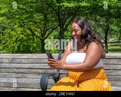 Eine afroamerikanische Frau auf ihrem Telefon, draußen auf einer Bank Stockfoto