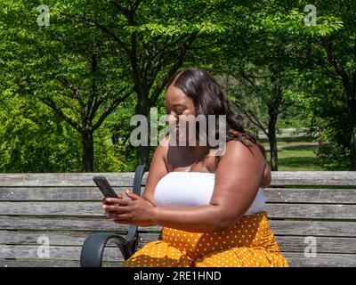 Eine afroamerikanische Frau auf ihrem Telefon, draußen auf einer Bank Stockfoto