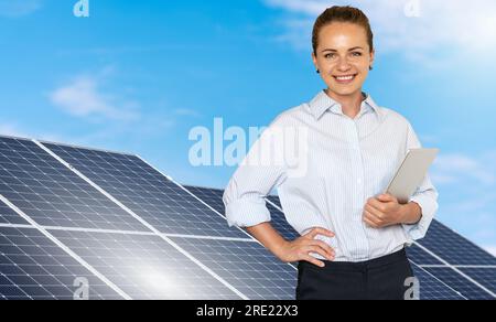 Junge Ingenieurin, die im Hintergrund einer Solarenergiefarm steht, ein digitales Tablet in der Hand hält und lächelt. Stockfoto