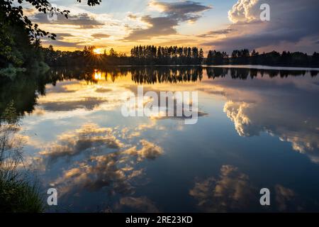 Ein See (Postfeldener Stausee) in Bayern im Naturschutzgebiet Höllbachtal bei Sonnenuntergang, der Himmel reflektiert im See. Deutschland. Stockfoto