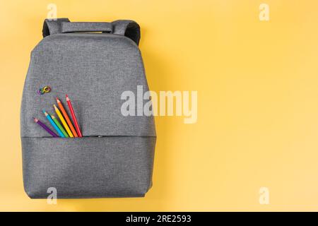 Grauer Rucksack mit unendlichem Autismus-Regenbogen-Symbol, farbige Bleistifte auf gelbem Hintergrund. Draufsicht. Welttag des Autismus, Autismusrechte Stockfoto